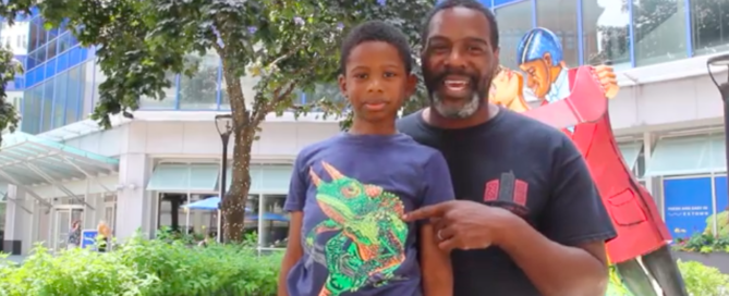 One older Black male wearing a blue tee shirt and his young son wearing a blue tee shirt with a lizard, standing in front of a sculpture talking about Sculptures and Stories.