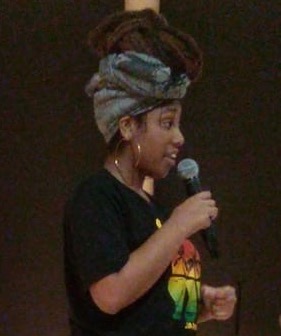 A young Black woman with hoop earrings, a head wrap, and a black Bob Marley t-shirt holds a microophone.