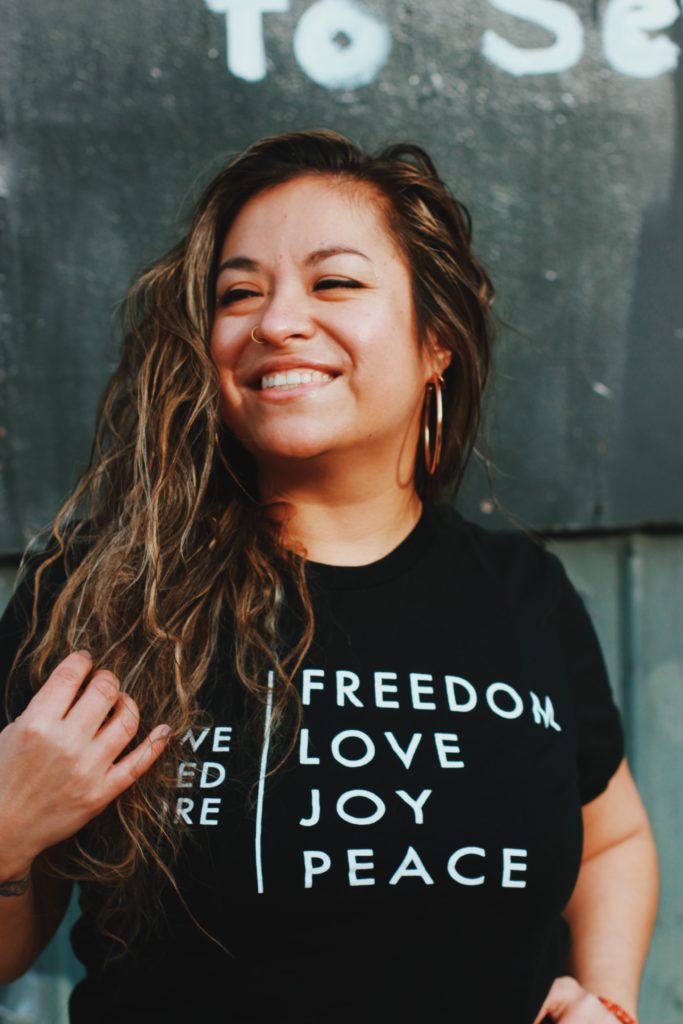 Nicole smiling, long hair tossed over one shoulder and wearing a shirt that says freedom love joy peace