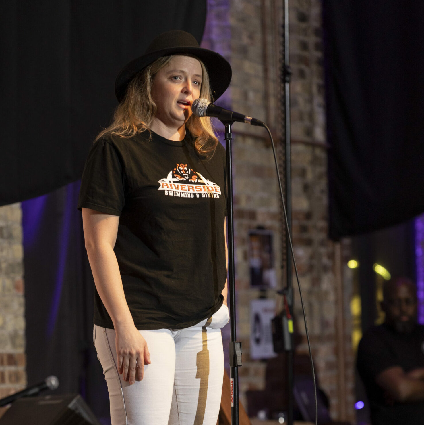 Ann Koller, a blonde white woman, speaks into a microphone on stage. She is wearing a black brimmed hat, black tshirt, and white pants.