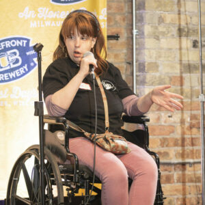 Jill Potkay, a red haired woman wearing an Ex Fabula shirt and sitting in a wheelchair, speaks into a microphone. She holds out one hand with the palm facing the crowd.