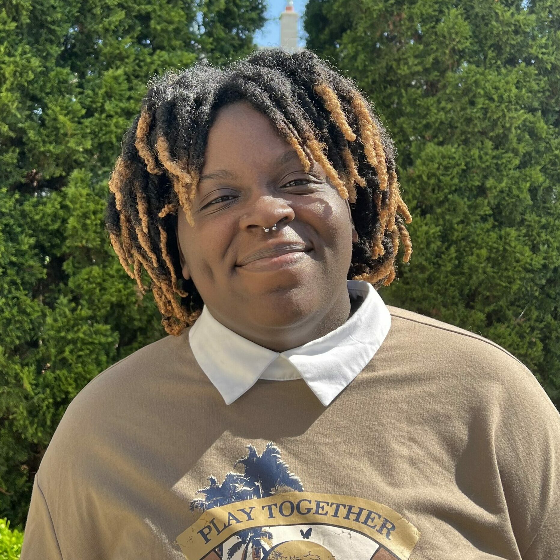 A person with dark skin and face-framing locs smiles. They are wearing a brown sweatshirt, and white collared shirt. There are trees in the background.