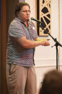 Eric, a white man with a patterned shirt and kahki shorts on, stands at a microphone telling a story.