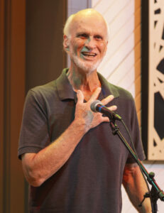 Jerry, an older white man with short facial hair and a grey shirt stands at the microphone telling a story. His hand is on his heart.