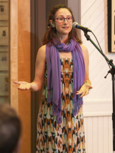 Sarah Beth, a middle-aged white woman is standing in a patterned dress and purple scarf at the microphone telling a story. Her hands are gesturing out and to her right.