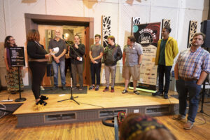 All of the storytellers stand in a line on stage with the emcee Michaela, a young black woman, stands in front.