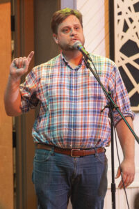 Paul, a middle-aged white man, is standing at a microphone telling a story. One hand is held up with his pointer finger pointing upwards.