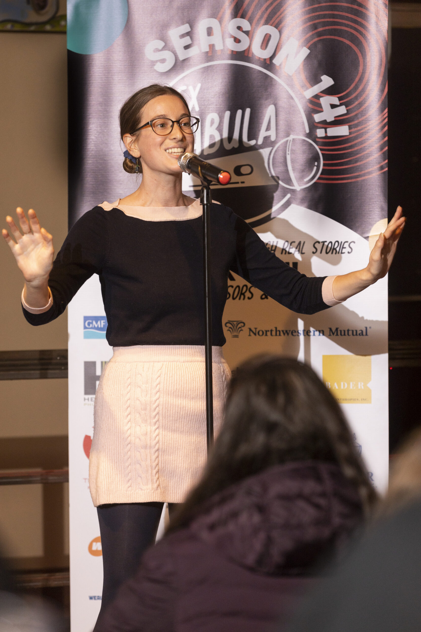 Anya, a white woman with her hair pulled into a low bun is wearing a black shirt and pink knit skirt. She holds out her arms and speaks into microphone