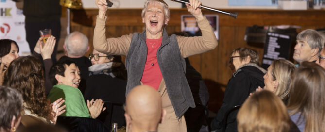 Woman in grey vest holds cane above her head and laughs