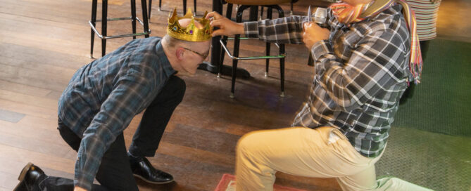 Joel, wearing a blue shirt and gold crown, kneels before Neil, the emcee wearing a plaid shirt and a scarf.