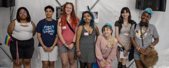 7 teens standing in a line, looking at the camera. They are of different genders and races and they are smiling. Many are wearing pride-themed bracelets and/or flags - including rainbow, pansexual, and nonbinary flags.