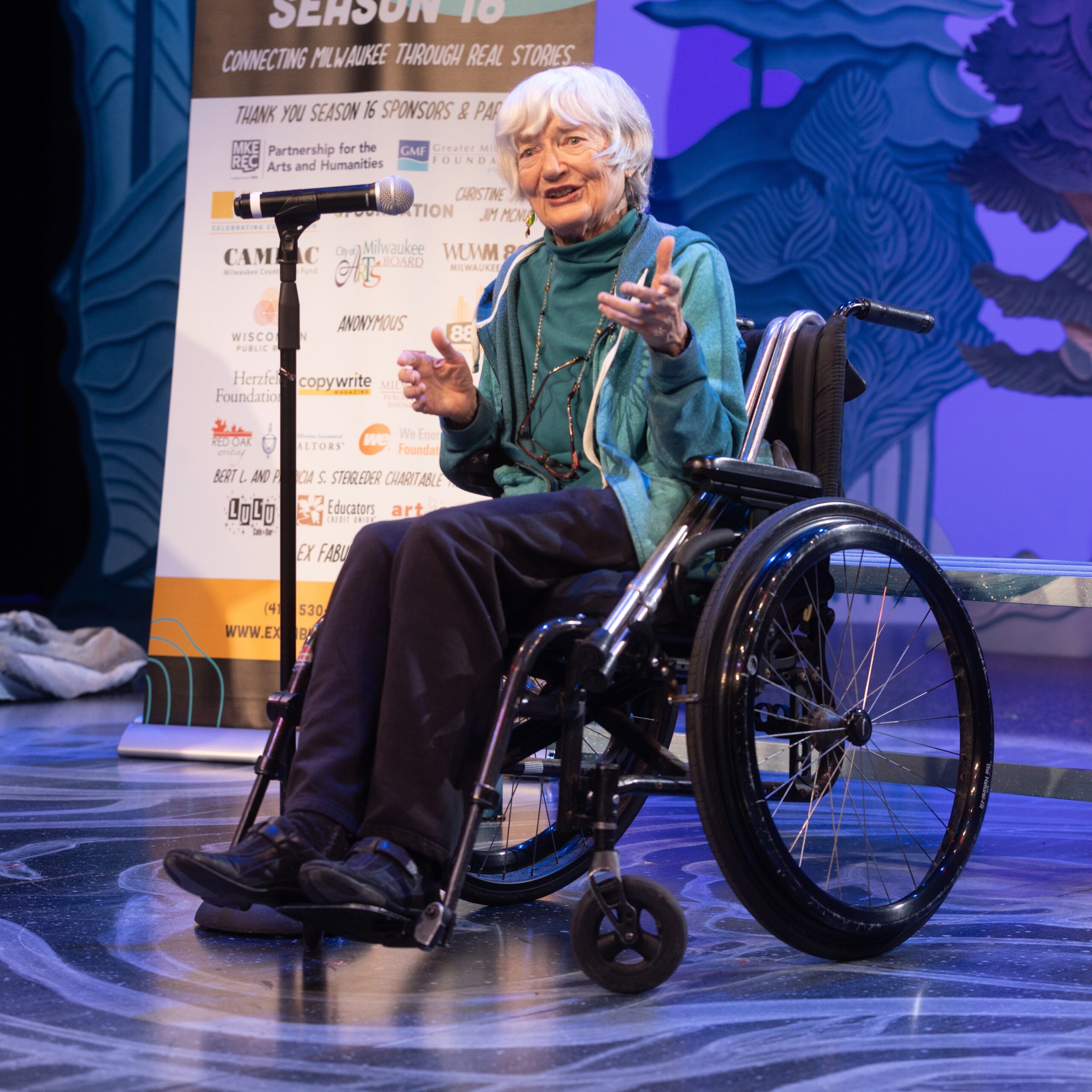 241203ExFabula_0153 Person in a wheelchair, on stage at an Ex Fabula event, speaking into a microphone on a stand.