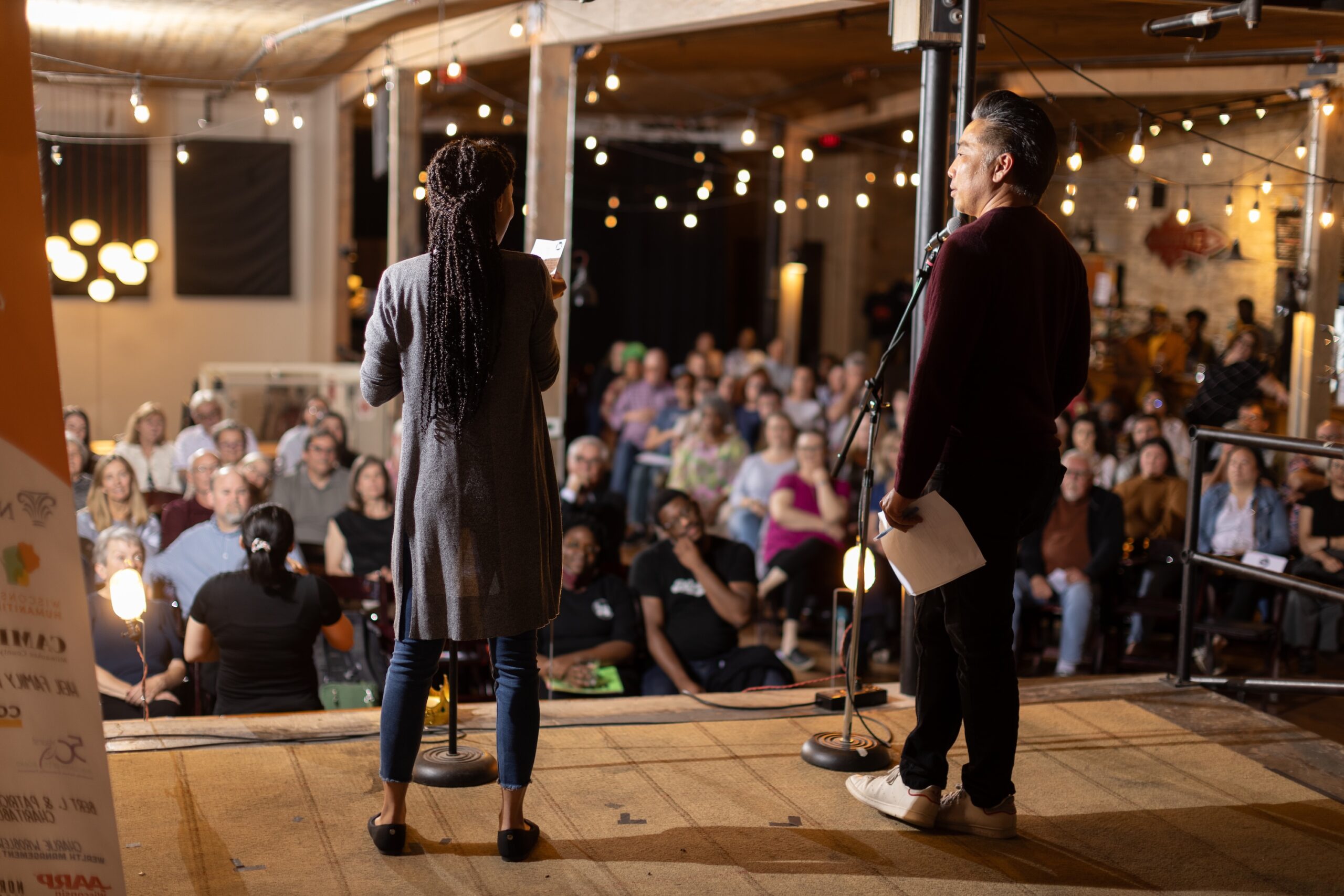 Two individuals on a stage, with their backs towards the camera, facing an audience