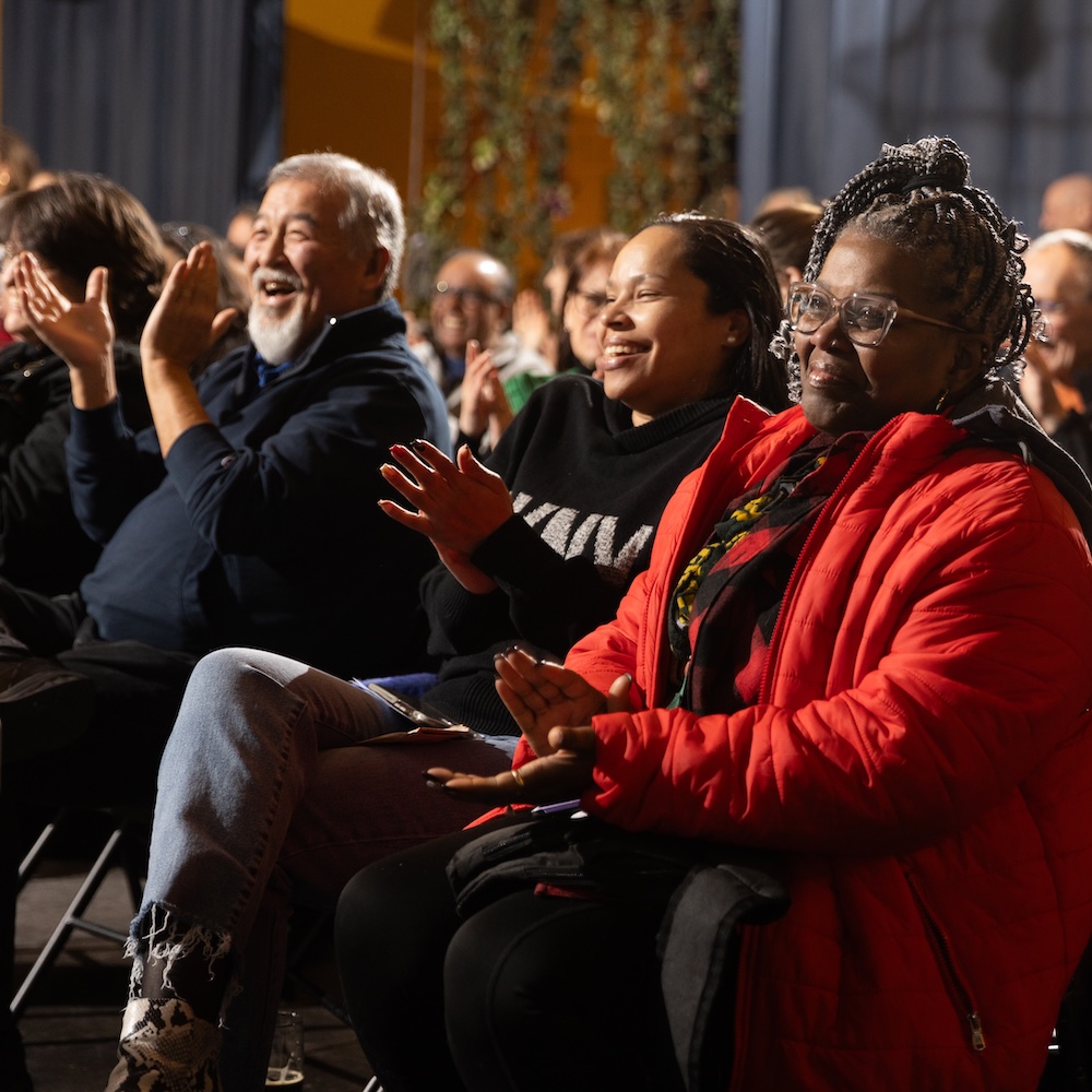 Photo of audience members applauding. Three individuals are in focus, smiling and clapping