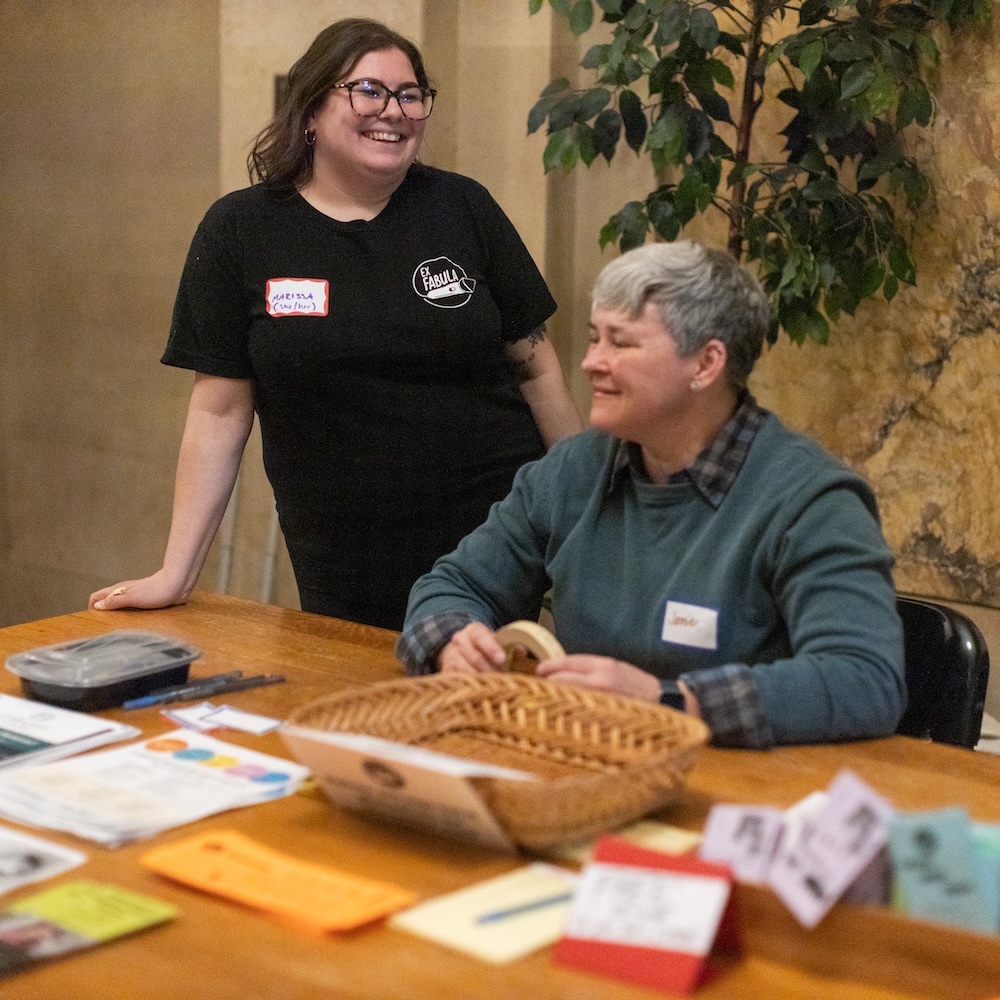 Photo of two volunteers for Ex Fabula, one sitting at a table with flyers and a basket, and one standing behind. 