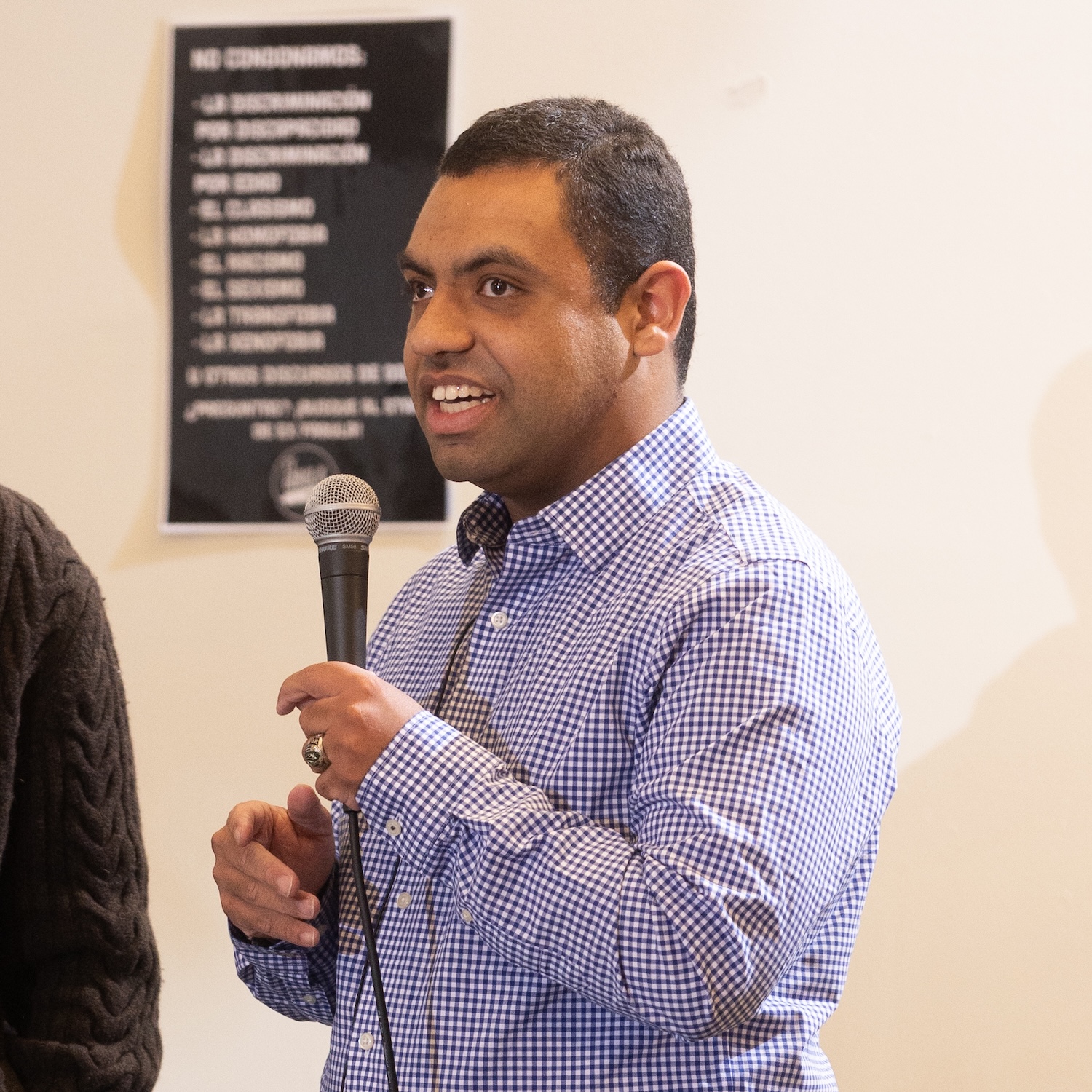 A man in his 20s holds a microphone while telling his story. He is blind, has light brown skin and short hair, and is wearing a navy and white checked button down shirt.