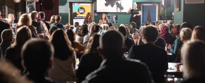 Photograph from the viewpoint of the back of audience at The Sugar Maple looking up at two emcees and one ASL interpreter.
