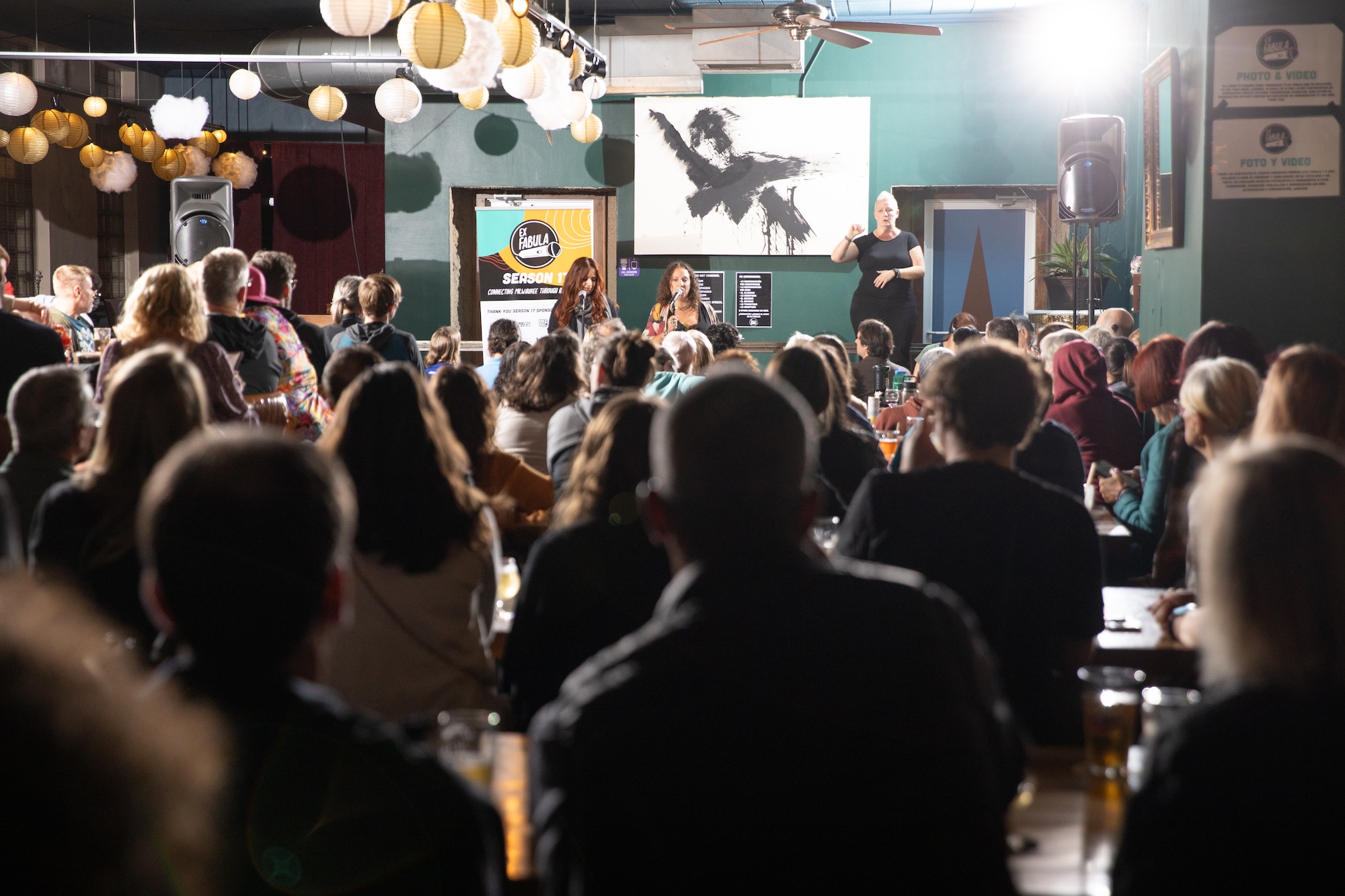 Photograph from the viewpoint of the back of audience at The Sugar Maple looking up at two emcees and one ASL interpreter.