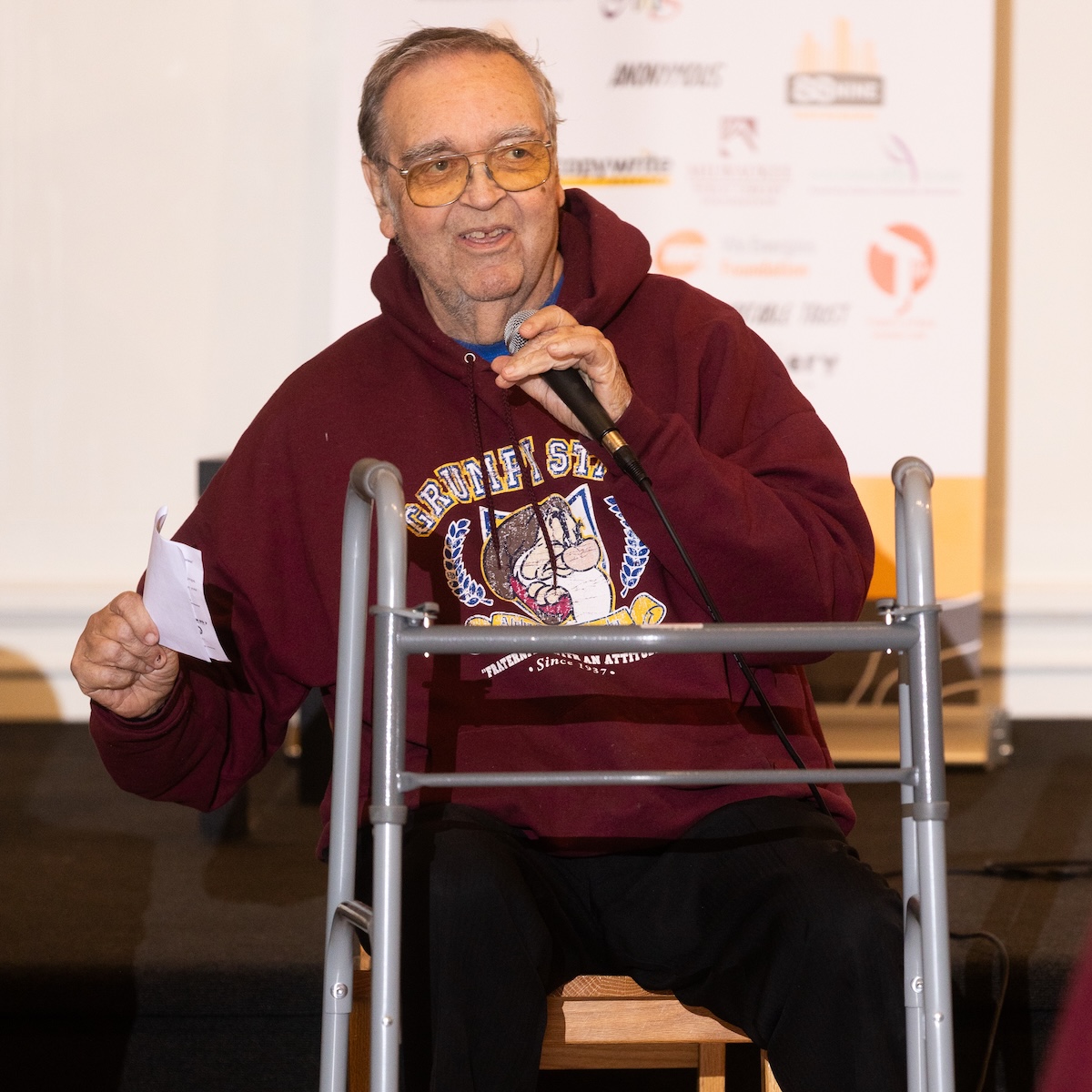 An older white man sits in chair with his walker in front of him. He holds a microphone and tells a story. He is smiling. He has tinted glasses and wears a maroon sweatshirt and black pants.