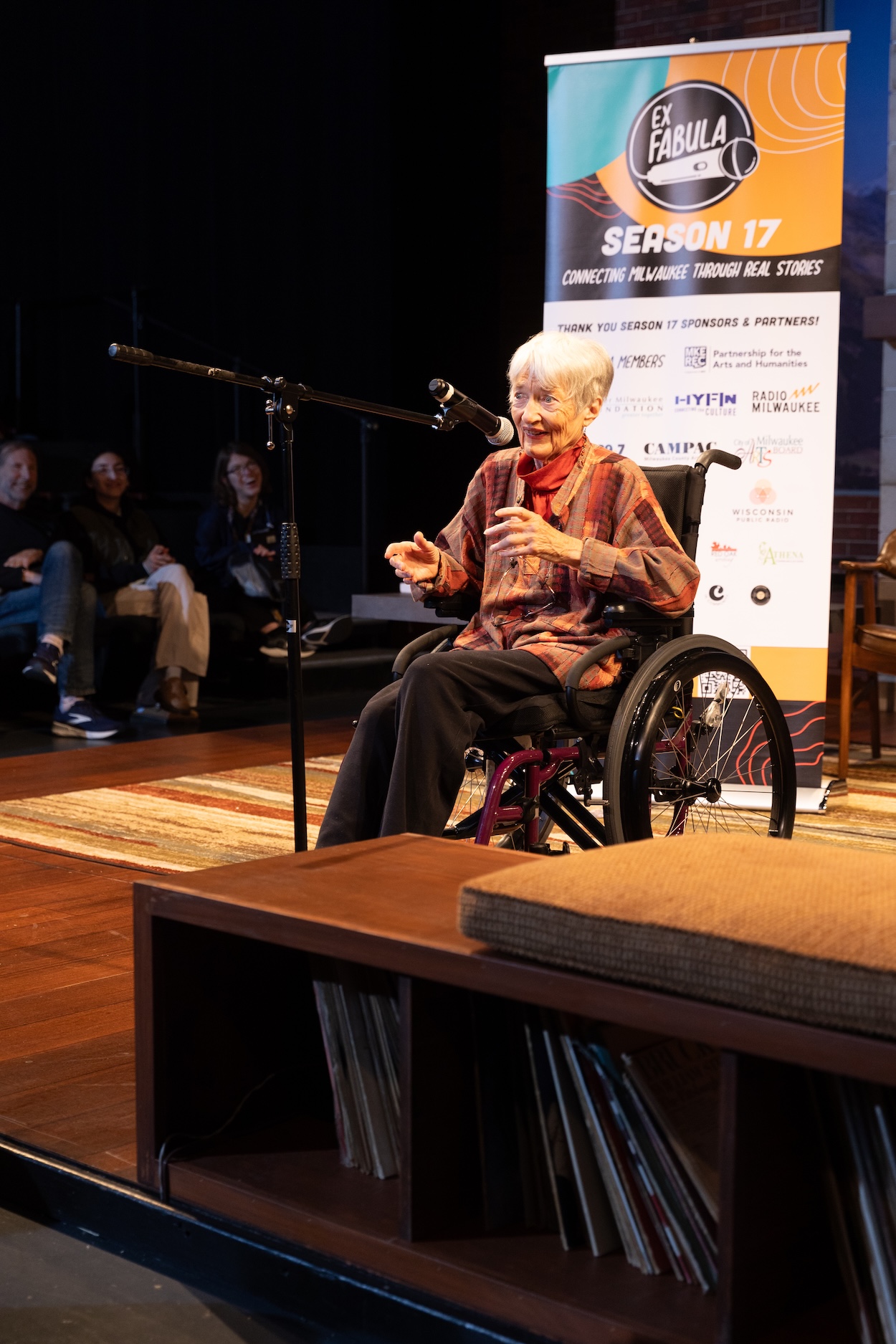 Person in a wheelchair, seated on a stage, speaking into a microphone.