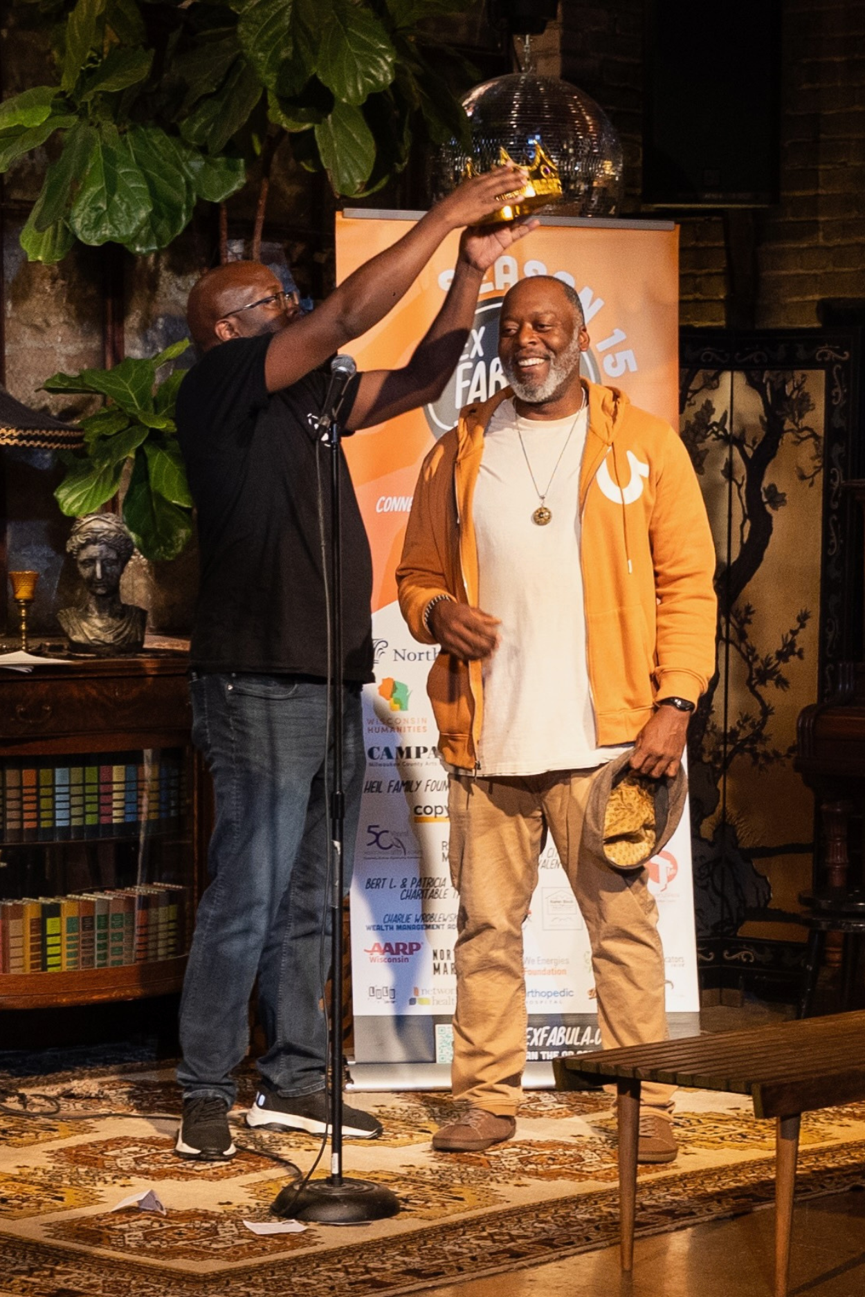 Two people behind a microphone. The person on the left is putting a plastic crown on the head of the person on the right.