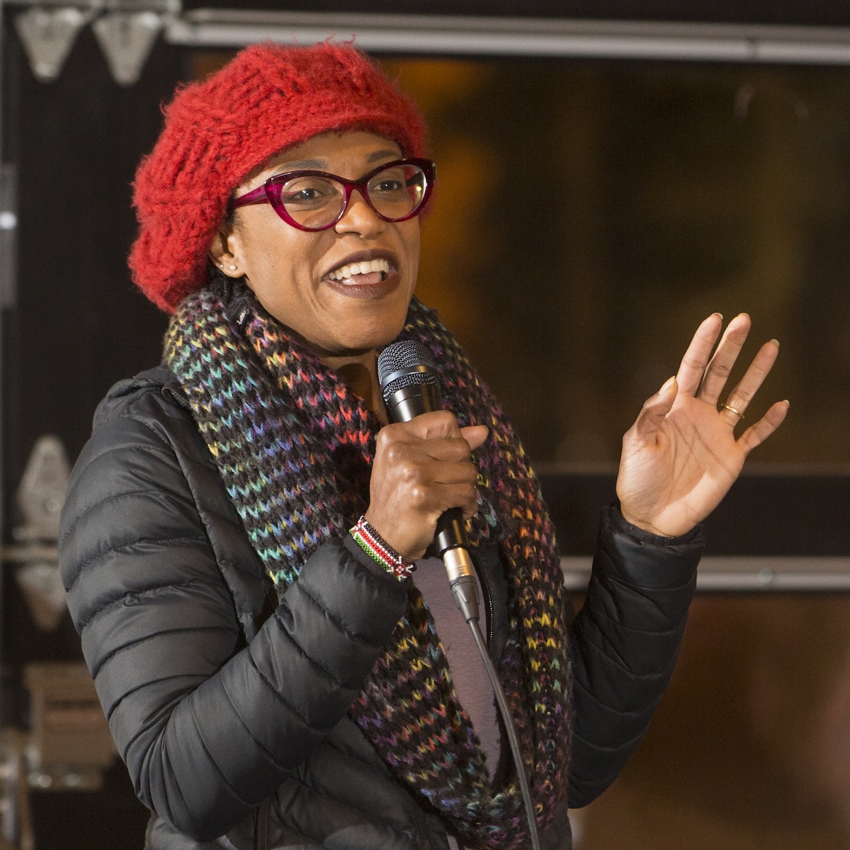Woman with dark skin, red glasses, and a red beret, holding a microphone and speaking. She is gesturing with her left hand and smiling. 