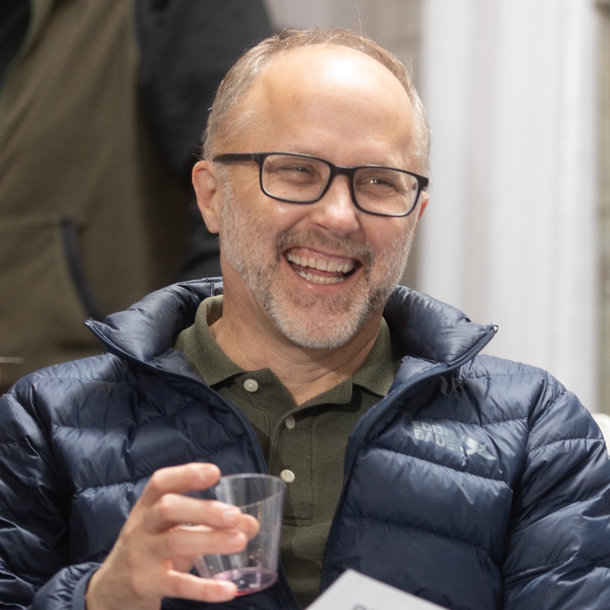 A man with light skin, short gray hair, and black glasses, and short facial hair. He's laughing and holding a drink and watching the stage at a StorySlam.