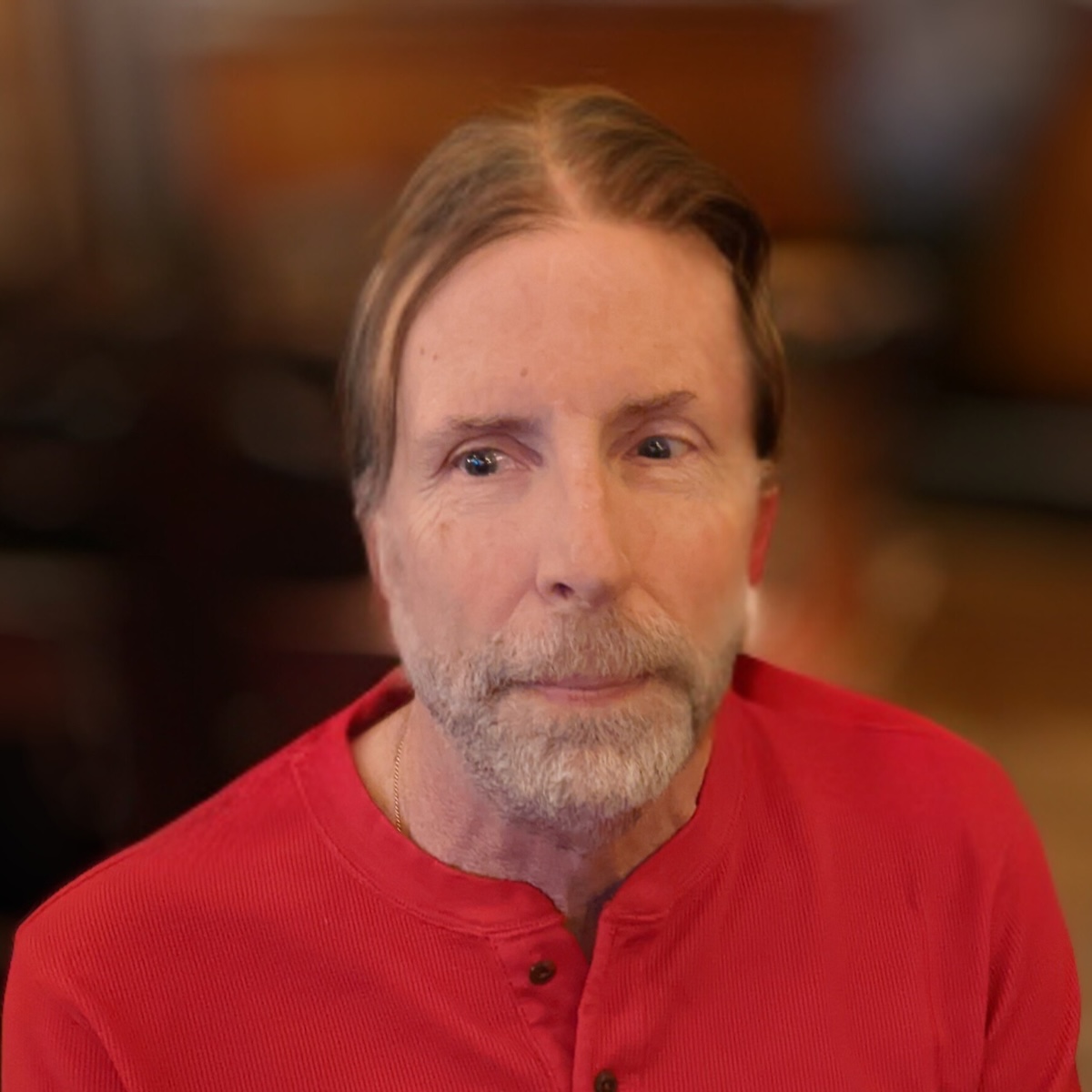 a person with light skin, light brown hair in a long ponytail, and a graying beard. They have a neutral look on their face. 