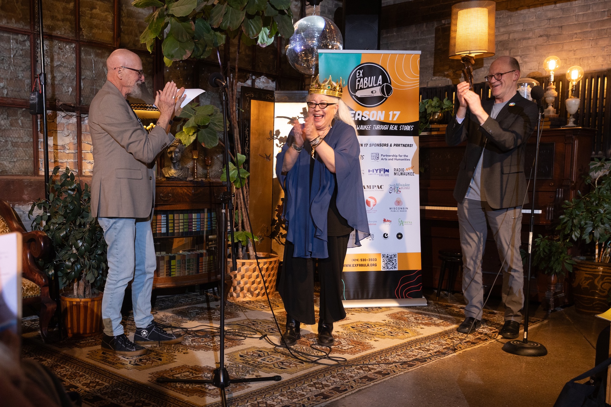 Photo of StorySlam: Growing Pains audience favorite Tracy Stockwell wearing her crown on stage, flanked by emcees Mark Weinberg and Joel Dresang