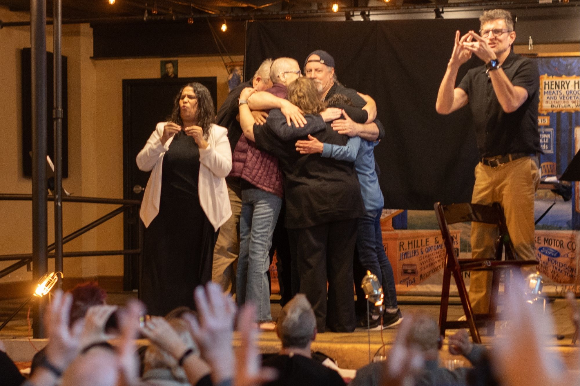 Storytellers from the seventh annual Deaf StorySlam: Jaw Dropping embrace in a group hug while audience members wave their hands in applause.