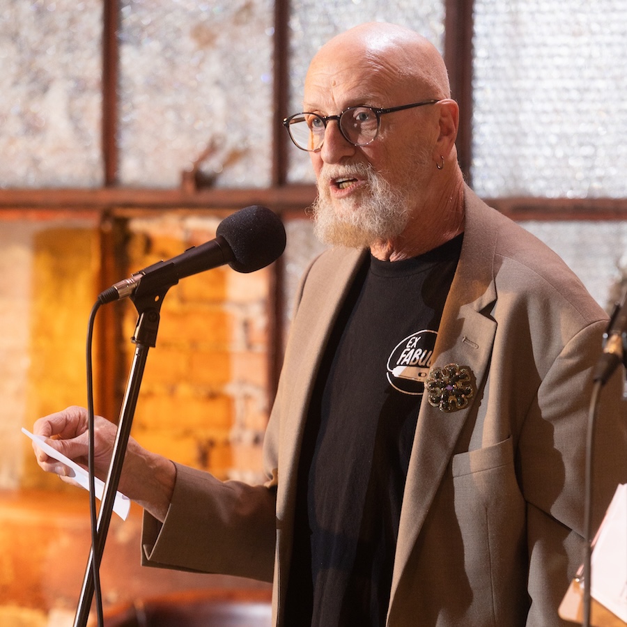 Photo of Mark Weinberg, a White man wearing a black Ex Fabula t-shirt under a tan blazer, on stage at an Ex Fabula event