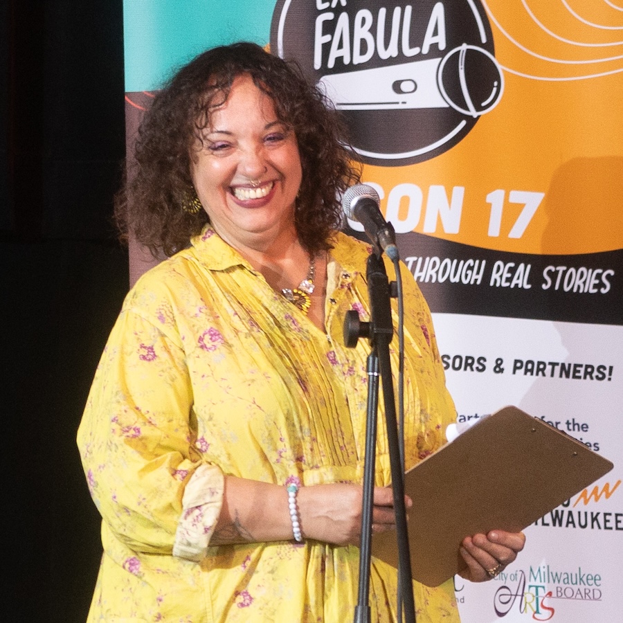 Photo of Shannon Sloan-Spice. She has short curly brown hair, is wearing bright yellow, and is standing behind a microphone on stage at an Ex Fabula event.