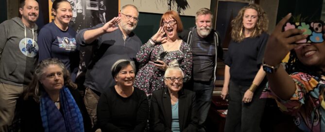 A group of adults gathered together in 2 rows with some sitting. They are smiling and a few are playfully posing for the camera. To the right of the photo, we can see a woman standing a few feet in front, taking an "us-ie" photo on her phone.